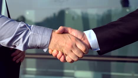 Close-up-of-successful-businessmen-greeting-each-other-near-office-building.-Two-young-colleagues-meeting-and-shaking-hands-in-urban-environment.-Handshake-of-business-partners-outdoor.-Side-view