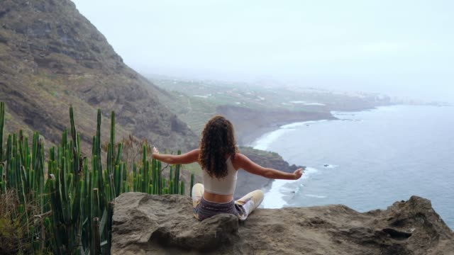 Young-woman-doing-yoga-in-the-mountains-on-an-island-overlooking-the-ocean-sitting-on-a-rock-on-top-of-a-mountain-meditating-in-Lotus-position