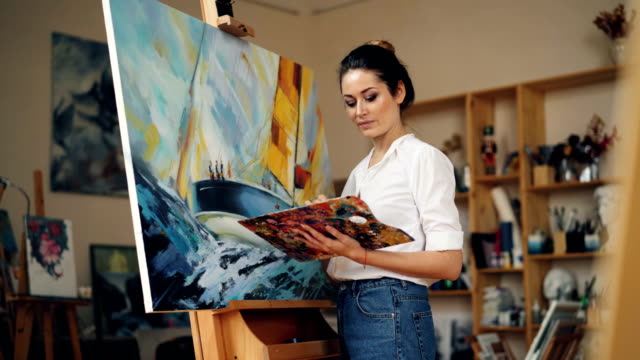Skillful-female-painter-is-working-in-studio-alone-painting-picture-on-easel-using-oil-paints,-palette-and-brush-enjoying-her-occupation.-People-and-work-concept.