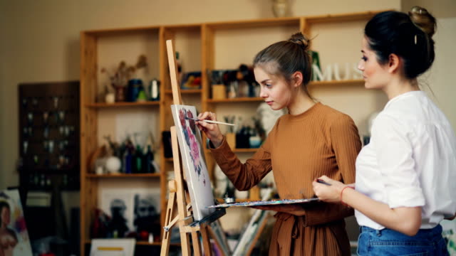 Talented-student-of-art-school-is-painting-picture-with-her-teacher-working-together-in-studio-full-of-artworks-and-tools.-Youth,-creativity-and-people-concept.