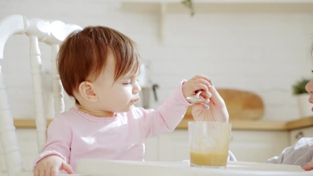 Side-view-of-cute-baby-girl-sitting-in-highchair-and-eating-with-spoon-vegetable-puree-helped-by-her-unrecognizable-mother