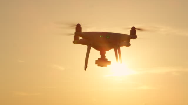SLOW-MOTION-SILHOUETTE:-Quadcopter-drone-flying-towards-the-sun-at-golden-sunset