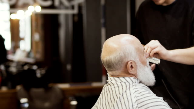Stylist-cuts-gray-beard-with-electric-razor-to-senior-man