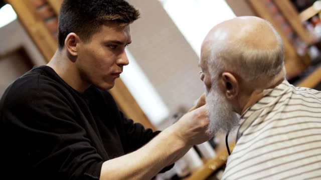 Stylist-cuts-beard-of-senior-man-with-electric-razor