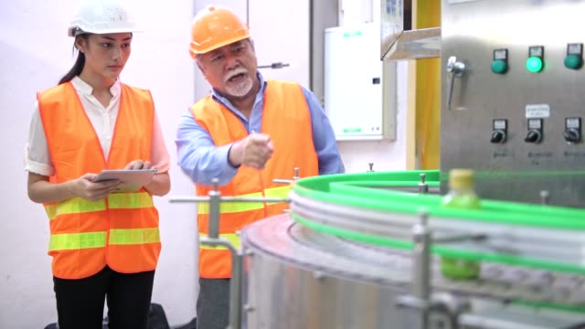 Senior-quality-inspector-teaching-junior-team-member-in-factory.-Chinese-old-male-with-his-young-team-discussing-quality-issue-with-bottle-production-line-in-background.