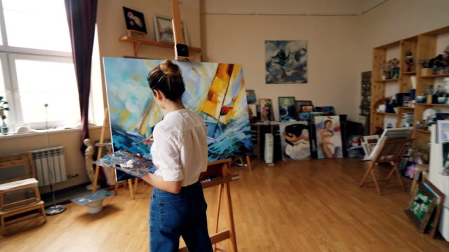 Pan-shot-of-serious-girl-professional-painter-working-in-studio-painting-marine-landscape-with-tempera-paints-holding-palette-and-brush.-Artworks-and-creativity-concept.