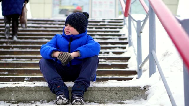 Boy-teenager-in-blue-down-jacket-lost-in-the-city.-He-sits-on-a-cold-staircase,-looks-around.