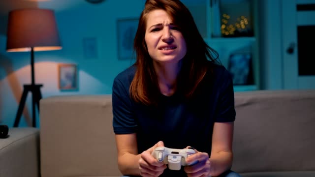 Sad-girl-loses-the-video-game-at-home-at-night.-Excited-gamer-woman-sitting-on-a-couch,-playing-and-losing-in-video-games-on-a-console,-using-a-wireless-controller.-Cozy-room,-lovely-home-atmosphere