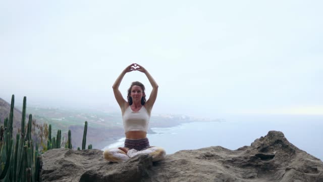A-woman-sitting-on-top-of-a-mountain-meditates-and-makes-a-gesture-with-her-hands-Maha-Sakal-.-Against-the-ocean-and-green-mountains