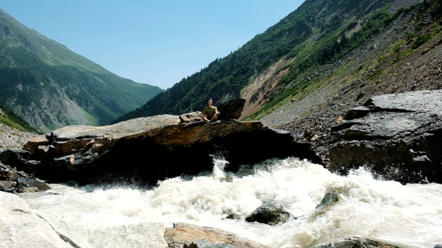 Man-sits-in-a-lotus-position-and-meditates-in-mountains.-River-flows-slow-motion