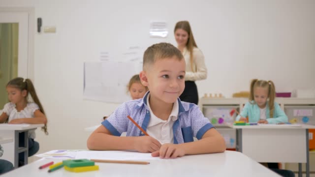 primary-school-children-draw-on-paper-with-colored-pencils-at-a-desk-with-young-teacher-in-light-classroom