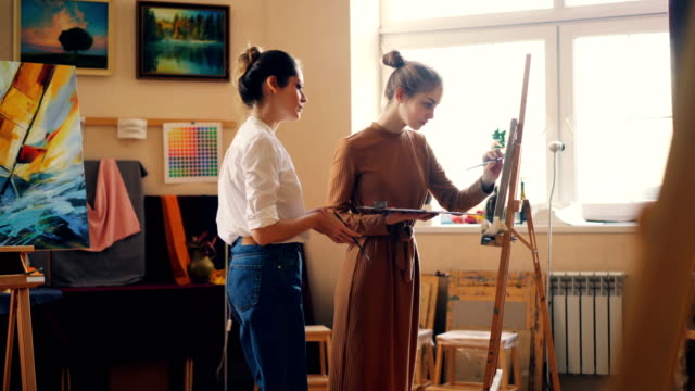 Attractive-girl-student-is-painting-during-art-class-in-studio,-her-teacher-is-coming-and-helping-her-teaching-giving-advice.-Cozy-workshop-with-artworks-and-tools-is-visible.