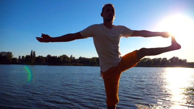 Young-man-standing-at-yoga-pose-on-wooden-jetty-at-lake.-Athlete-balancing-on-one-leg-at-nature.-Sporty-guy-doing-stretch-exercise-outdoor.-Concept-of-healthy-active-lifestyle.-Slow-motion-Close-up
