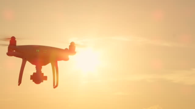 SLOW-MOTION-CLOSE-UP-LENS-FLARE:-Small-filming-drone-flying-on-golden-sunset-sky