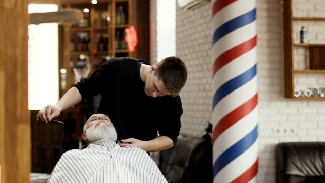 Professional-barber-cuts-beard-of-senior-man-in-barbershop