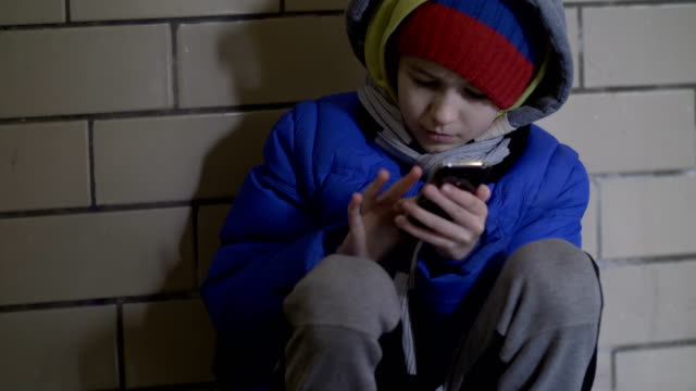 armer-Junge-sitzt-auf-dem-Boden-auf-der-Straße-am-Abend-und-nutzt-das-Telefon-im-winter