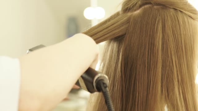 Hairstylist-combing-long-hair-and-straightening-with-hair-tongs-in-hairdressing-salon.-Close-up-female-hairdresser-straightening-hair-during-hairstyling-in-beauty-salon