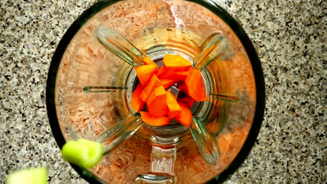 Stalk-of-an-apium-fall-on-carrots-in-a-blender-bowl.-Slow-motion.	Shooting-in-kitchen.-Top-view.