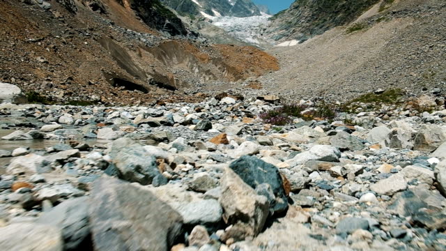 Steinigen-Moräne-in-den-Bergen-auf-dem-Hintergrund-des-Gletschers,-Slow-Motion.-Gebirgsbach-und-Wildblumen,-Bewegung-der-Kamera
