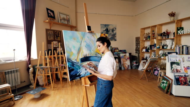 Serious-artist-is-working-in-workroom-standing-in-front-of-easel-with-palette-and-brush-and-painting-picture-boat-and-sea-waves-in-acrylic-paints.-People-and-hobby-concept.