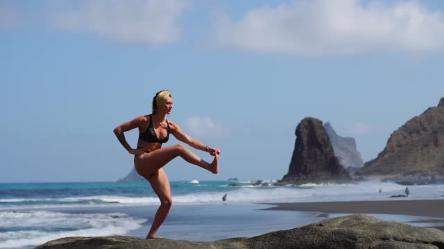 Young-girl-in-bikini-balancing-standing-on-one-leg-doing-yoga-standing-on-a-rock-by-the-ocean-on-a-black-sand-beach