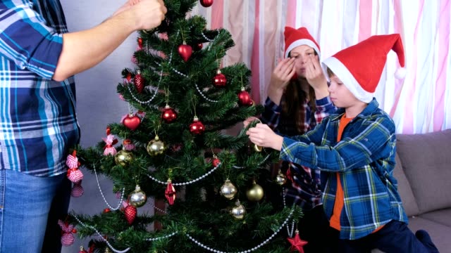 Family-dad,-mom-and-son-in-Christmas-hats-decorate-Christmas-tree-with-beads-and-balls.