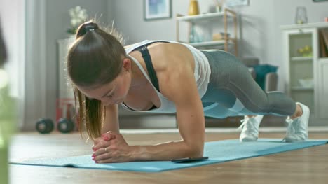 Strong-Beautiful-Fitness-Girl-in-Athletic-Workout-Clothes-is-Doing-a-Plank-Exercise-While-Using-a-Stopwatch-on-Her-Phone.-She-is-Training-at-Home-in-Her-Living-Room-with-Cozy-Interior.
