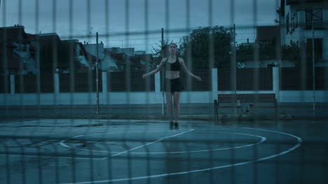 Beautiful-Energetic-Fitness-Girl-in-Black-Athletic-Top-and-Shorts-is-Skipping/Jumping-Rope.-She-is-Doing-a-Workout-in-a-Fenced-Outdoor-Basketball-Court.-View-from-Behind-the-Fence.-Evening-After-Rain.