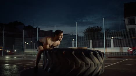Strong-Muscular-Fit-Young-Shirtless-Man-is-Doing-Exercises-in-a-Fenced-Outdoor-Basketball-Court.-He's-Flipping-a-Big-Heavy-Tire-in-an-Afternoon-After-Rain-in-a-Residential-Neighborhood-Area.