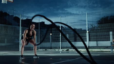 Schöne-energetische-Fitness-Girl-Übungen-mit-Battle-Ropes.-Sie-ist-ein-Training-in-einem-eingezäunten-Basketballfeld-im-freien-tun.-Abend-Footage-nach-Regen-in-einer-Wohngegend.