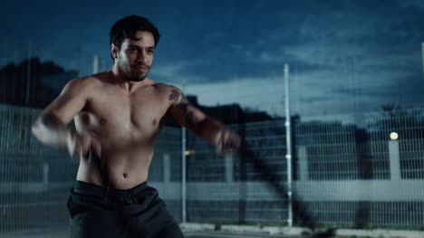 Strong-Muscular-Fit-Shirtless-Young-Man-is-Doing-Exercises-with-Battle-Ropes.-He-is-Doing-a-Workout-in-a-Fenced-Outdoor-Basketball-Court.-Evening-Footage-After-Rain-in-a-Residential-Neighborhood-Area.