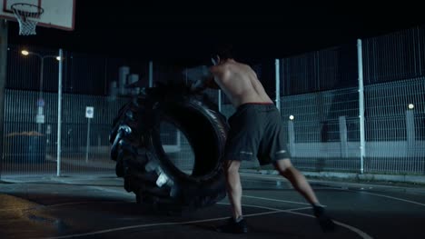 Backshot-of-a-Strong-Muscular-Fit-Young-Shirtless-Man-Doing-Exercises-in-a-Fenced-Outdoor-Basketball-Court.-He's-Flipping-a-Big-Heavy-Tire-in-a-Night-After-Rain-in-a-Residential-Neighborhood-Area.