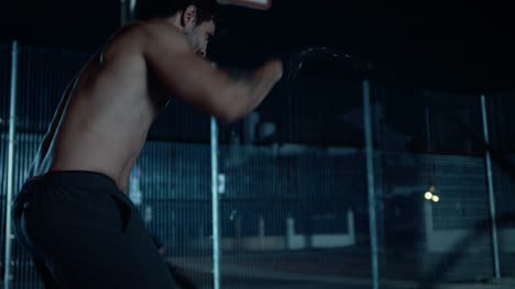 Backshot-of-a-Strong-Muscular-Fit-Shirtless-Young-Man-Doing-Exercises-with-Battle-Ropes.-He-is-Doing-a-Workout-in-a-Fenced-Outdoor-Basketball-Court.-Night-Footage-After-Rain-in-a-Residential-Neighborhood-Area.