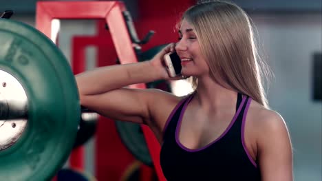 Lächelnde-Pass-Frau-im-Gespräch-am-Telefon-im-Fitnessstudio-Gewichtsraum