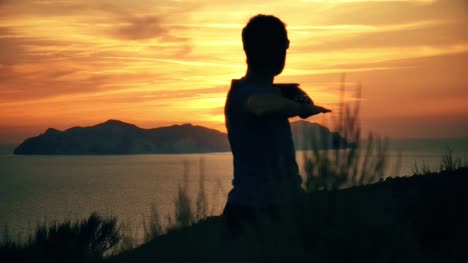 Silhouette-eines-Mannes-erstreckt-sich-auf-einem-Berg-auf-einer-Insel-bei-Sonnenuntergang-mit-dem-Meer-im-Hintergrund