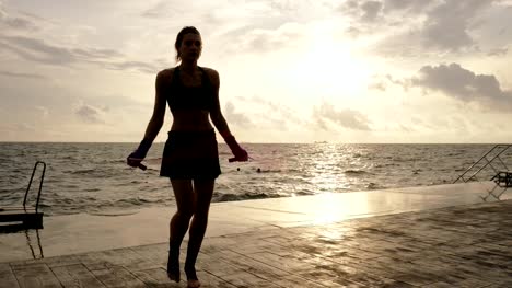 Young-woman-working-out-on-the-jump-rope-against-the-sun-by-the-beach-in-slowmotion.-Lens-flare.-Girl-jumping-on-a-skipping-rope-by-the-sea.-Shot-in-4k