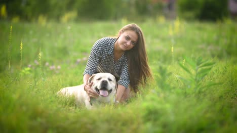 pretty-woman-is-stroking-her-big-white-labrador-and-looking-in-camera-in-a-middle-of-a-green-field,-green-grass-around