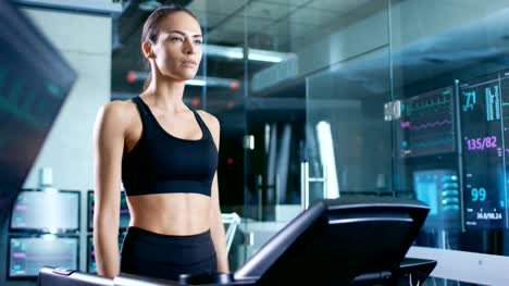 Beautiful-Woman-Athlete-Wearing-Sport-Bra,-Walks-on-a-Treadmill-in-a-Sports-Science-Laboratory.-In-the-Background-Monitors-Show-EKG-Data-and-Health-Data.