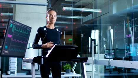 In-Scientific-Sports-Laboratory-Beautiful-Woman-Athlete-Runs-on-a-Treadmill-with-Electrodes-Attached-to-Her-Body,-Monitors-Show-EKG-Data-on-Display.-Slow-Motion.