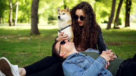 Cheerful-young-man-is-lying-on-the-grass-in-the-park-with-his-head-on-his-wife's-legs-while-attractive-smiling-woman-is-talking-to-him-and-caressing-well-bred-dog.