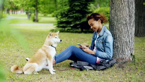 Seitenansicht-des-ziemlich-Mischlinge-Mädchen-mit-Smartphone-im-Park-unter-Baum-entspannen,-während-ihre-süße-Shiba-Inu-Hund-in-der-Nähe-von-ihrer-Besitzerin-sitzt-und-die-Natur-genießen.