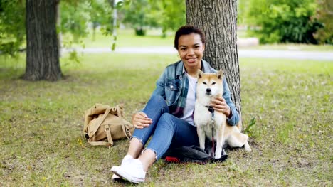 Portrait-of-African-American-girl-loving-dog-owner-sitting-in-park-on-grass-with-her-beautiful-pet,-smiling-and-looking-at-camera.-Loving-animals-and-nature-concept.