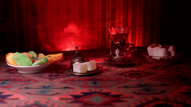 Arabian-tea-in-vintage-glass-with-eastern-snacks-on-a-carpet.-Eastern-tea-ceremony-on-dark-background-with-lights-and-smoke.-Empty-space.-Selective-focus.-Slider-shot