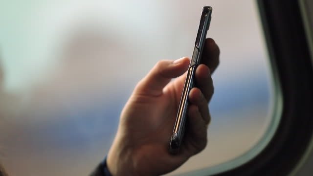 Close-up-of-hands-holding-smartphone-device-with-blurred-background-landscape-in-motion.-Person-surfing-the-internet-and-checking-emails-while-commuting-by-train