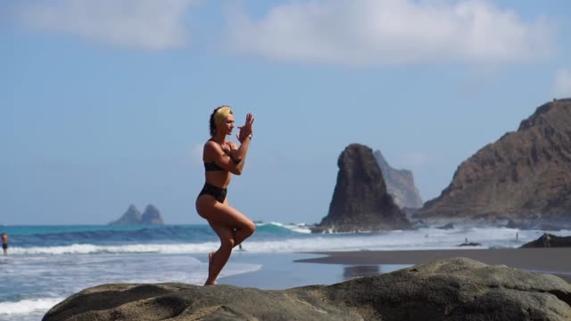 Junges-Mädchen-im-Bikini,-balancieren-auf-einem-Bein-tut-Yoga-steht-auf-einem-Felsen-am-Meer-auf-einem-schwarzen-Sandstrand