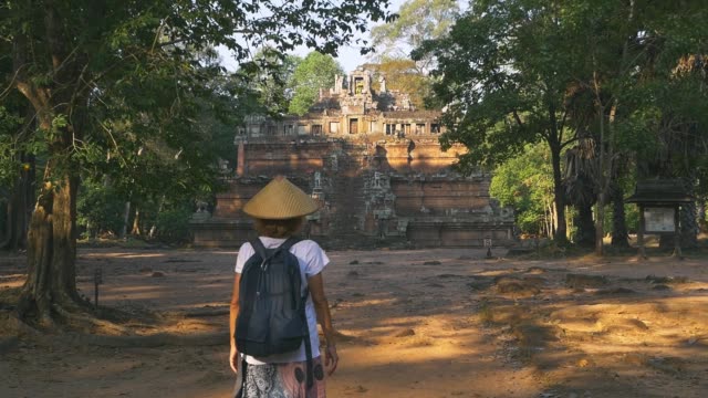 Slow-motion:-one-tourist-visiting-Angkor-ruins-at-sunrise,-travel-destination-Cambodia.-Woman-with-traditional-hat-walking-to-Baphuon-temple-in-the-jungle,-rear-view.