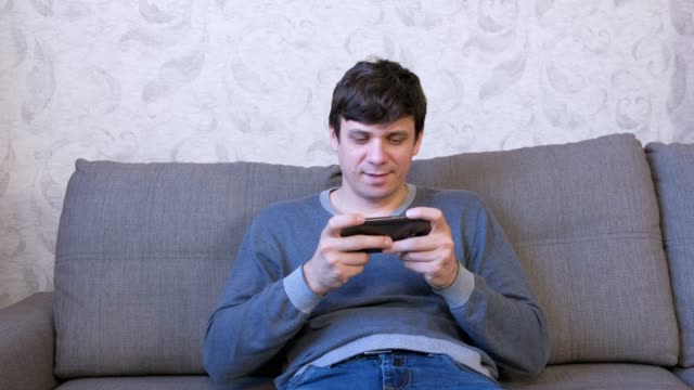El-hombre-está-jugando-al-juego-en-su-teléfono-móvil-sentado-en-el-sofá.