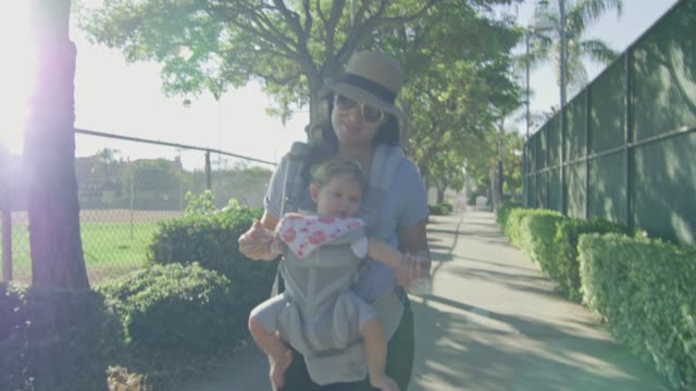 Woman-smiling-as-she-walks-with-baby-in-carrier-at-a-park