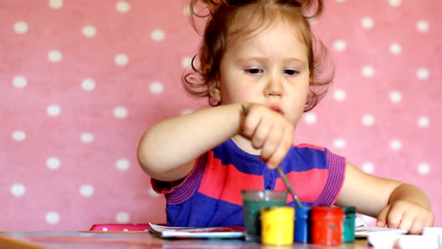 Lustige-kleine-Mädchen-zeichnen-Gouache-an-einem-Tisch-auf-einem-rosa-Hintergrund.-Porträt-von-einem-niedlichen-Baby-mit-einem-Pinsel-und-farbige-Lacke.-Kleine-Künstler.-Kreatives-Konzept.-Kindheit.-Vorschulkind-Bildung