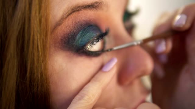 Close-up-of-young-female-model-is-being-maked-up-in-studio.-Artist-is-applying-black-liner-to-lower-lid-of-the-right-eye-using-a-brush.-Side-view